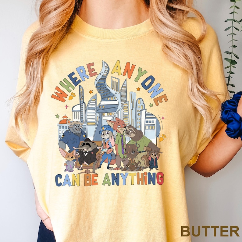 May include: Yellow t-shirt with a colorful graphic featuring cartoon characters and a cityscape. The text on the shirt reads "WHERE ANYONE CAN BE ANYTHING". The shirt is made of a soft material.