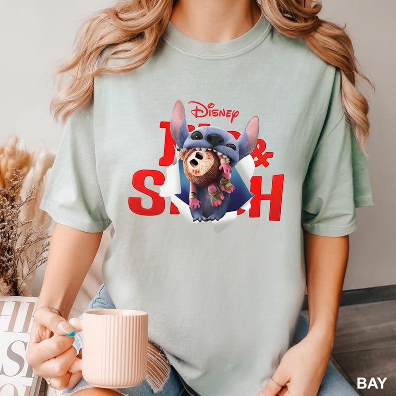 Puede incluir: Camiseta verde claro con un dise&ntilde;o de Stitch de Disney y un koala marr&oacute;n. La palabra "Disney" est&aacute; encima de las palabras rojas "Stitch &" en la camiseta. El dise&ntilde;o muestra a Stitch rompiendo un agujero en la tela.