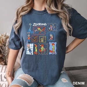 May include: A denim blue t-shirt featuring the word "Zootopia" above a grid of cartoon character illustrations. The shirt has a crew neck and short sleeves. The word "DENIM" is printed in white at the bottom right.