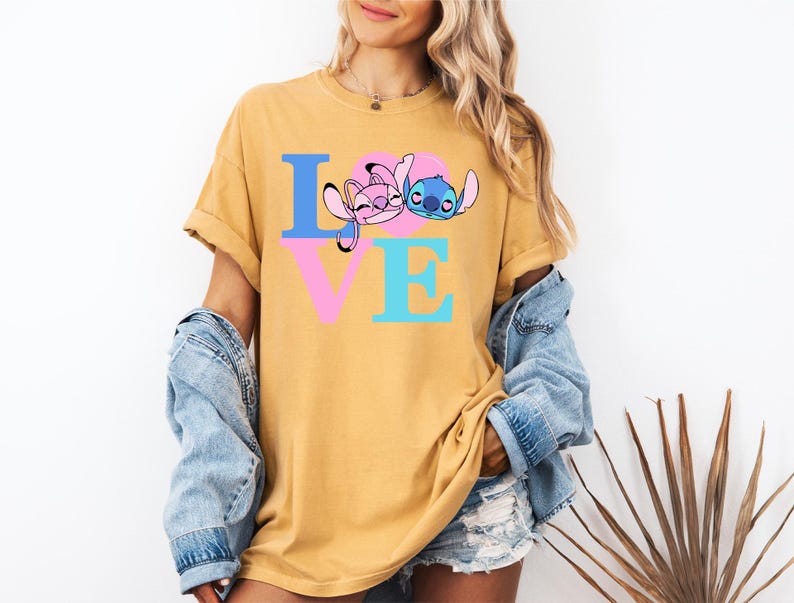 May include: Mustard yellow t-shirt with the word "LOVE" in blue, pink, and turquoise. The "O" is replaced with a cartoon image of Stitch and Angel. The shirt is worn with a denim jacket.