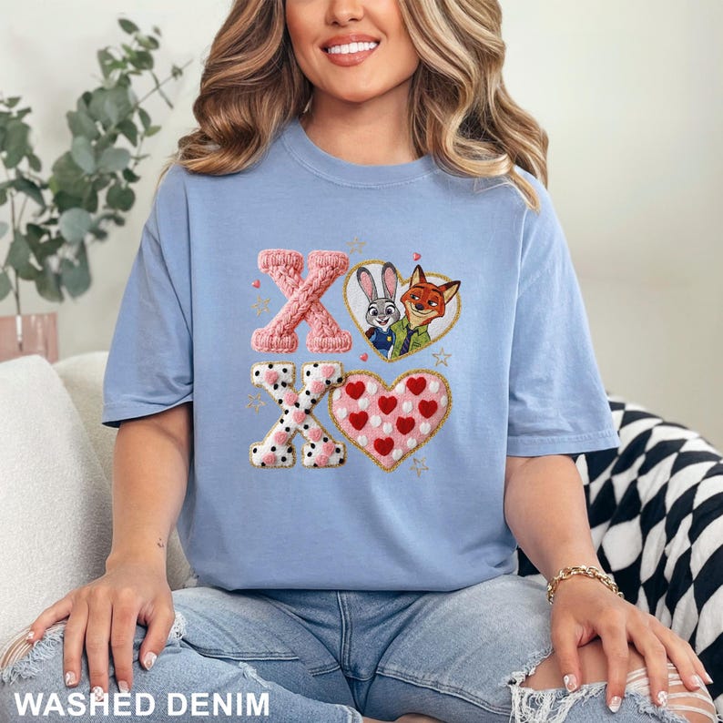 May include: A light blue t-shirt with an embroidered design featuring the characters Judy Hopps and Nick Wilde from Zootopia, along with the letters "XO" and heart shapes. The shirt is made of a soft material and is a casual style.