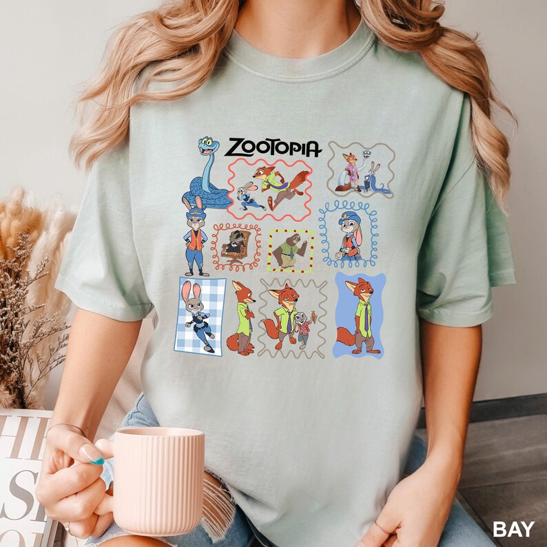 May include: A light green t-shirt featuring the word "Zootopia" above a grid of cartoon character illustrations. The shirt has a crew neck and short sleeves. The illustrations include a snake, a fox, a bunny, and other characters from the movie.