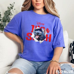 May include: A blue t-shirt featuring the Disney characters Lilo & Stitch. The graphic shows Stitch bursting through a torn paper effect. The text "Disney Lilo & Stitch" is printed in red. The shirt is a solid blue color.