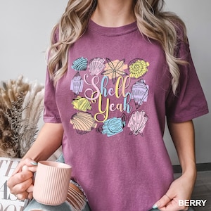 May include: Berry-colored t-shirt with a colorful seashell graphic and the text "Shell Yeah". The design includes various seashell illustrations in shades of blue, yellow, and pink. The shirt is made of a soft material.