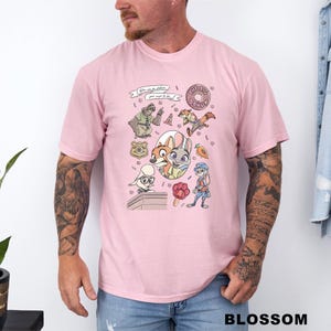 Puede incluir: Camiseta rosa claro con personajes de dibujos animados y el texto "You can be whatever you want to be" y "The Big Donut". La camiseta tiene cuello redondo y mangas cortas.