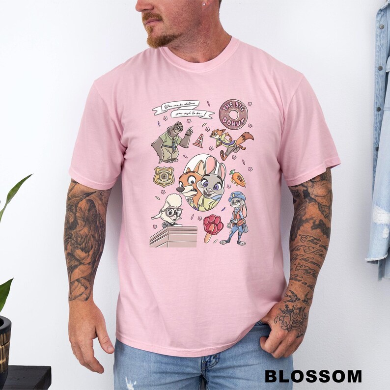 Puede incluir: Camiseta rosa claro con personajes de dibujos animados y el texto "You can be whatever you want to be" y "The Big Donut". La camiseta tiene cuello redondo y mangas cortas.