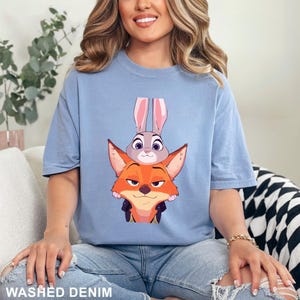 Puede incluir: Camiseta azul claro con un dise&ntilde;o de zorro y conejo de dibujos animados. El zorro es naranja con una chaqueta negra y el conejo es gris. El texto "WASHED DENIM" est&aacute; en la parte inferior de la imagen.