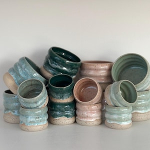 Könnte beinhalten: Eine Sammlung handgefertigter Keramikbecher in verschiedenen Farben, darunter Türkis, Grün und Rosa. Die Becher haben ein strukturiertes, geripptes Design und einen gesprenkelten, natürlich getönten Boden. Die Anordnung zeigt die Vielfalt der Größen und Farben.