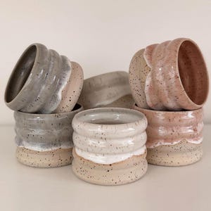 Könnte beinhalten: Eine Ansammlung von fünf Keramikbechern mit gerippter, zylindrischer Form. Jeder Becher hat eine gesprenkelte Basis und einen glatten, glasierten oberen Teil in Grau-, Creme- und Altrosatönen mit sichtbaren Tropfenmustern am Rand.