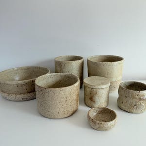 Op de afbeelding: Een verzameling van zeven keramische vaten in een gespikkelde beige afwerking, waaronder een brede ondiepe kom, verschillende cilindrische bekers van verschillende hoogtes, een kleine container met deksel en een piepklein rond schaaltje.