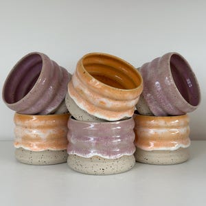 Könnte beinhalten: Eine Ansammlung von fünf Keramikbechern mit gerippter, wellenförmiger Textur. Die Becher haben eine gesprenkelte cremefarbene Basis mit glasierten oberen Abschnitten in sanften Rosa-, Lavendel- und warmen Orangetönen, gestapelt auf einer Fläche.