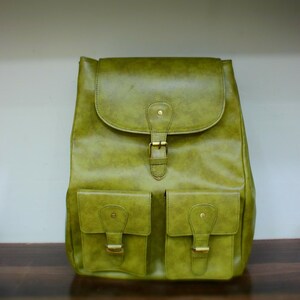 Puede incluir: Una mochila de cuero verde con cierre de solapa y dos bolsillos delanteros. La mochila presenta hebillas y herrajes de color latón. La bolsa está hecha de un material de cuero texturizado.
