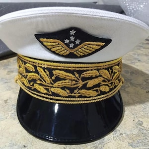 Puede incluir: Un sombrero blanco de estilo militar con una banda negra y bordados dorados. El sombrero tiene un emblema dorado con alas y cinco estrellas en la parte delantera.