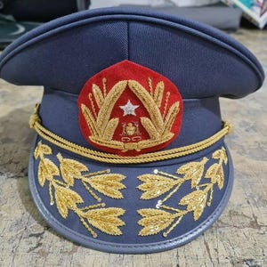 Pode incluir: Um chapéu de estilo militar azul escuro com guarnição dourada e um emblema bordado em vermelho e dourado com uma estrela e um brasão. O chapéu tem um desenho bordado em ouro ao redor da borda inferior.