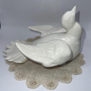 May include: A white ceramic dove figurine with outstretched wings, perched on a beige lace doily. The dove is smooth and glossy, with detailed feather impressions. The doily has a scalloped edge.