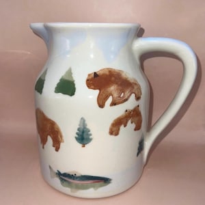 May include: A white ceramic pitcher featuring a hand-painted design of brown bears, green pine trees, and a single fish near the base. The pitcher has a curved handle on the right side and a pouring spout at the top rim, with a smooth glossy finish.