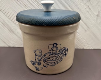 May include: A tan ceramic jar with a blue wooden lid and a white knob. The jar features a blue illustration of a teddy bear pulling a basket with a doll inside. A charming decorative piece.