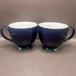 May include: Two navy blue ceramic coffee mugs with white interiors and green bases. Each mug has a rounded body, a curved handle, and a wide opening. The mugs are designed for hot beverages.
