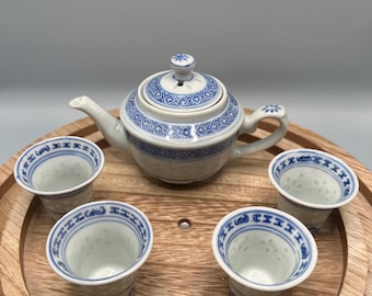 May include: A white porcelain tea set with blue floral patterns, including a teapot and four small cups. The set is arranged on a round wooden tray. The teapot has a lid and a handle. The cups have a flared shape.