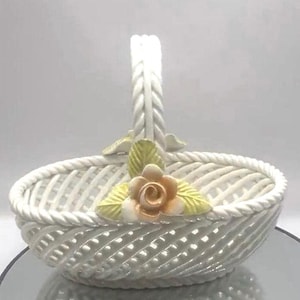 May include: A white ceramic basket featuring a woven lattice design and a twisted rope-style handle. The basket is decorated with a single peach-colored rose and two light green leaves attached to the base of the handle on the front side.