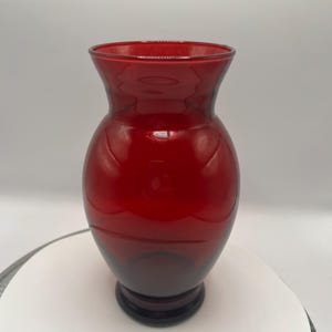 May include: A ruby red glass vase with a rounded body and flared rim. The vase has a dark base and a smooth, glossy finish. The vase is likely used for displaying flowers or as a decorative item.