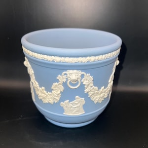 May include: A light blue ceramic planter with white relief detailing. The planter features a decorative rope-like border and white relief images of figures, floral garlands, and lion head ring handles. The planter is set against a dark background.