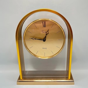 May include: An arched mantel clock with a gold-toned frame and a circular gold face. The clock features black hands, black hour markers, and the text Equity and Quartz printed on the face. The design includes a clear glass panel within the frame.
