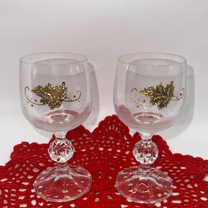 May include: Two clear wine glasses with gold leaf designs and crystal-like stems. The glasses sit on a red, open-weave doily. The gold leaf design includes a leaf and curved lines with small red dots.