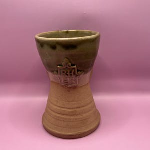 May include: A handmade ceramic cup with a unique hourglass shape. The top is a dark green, transitioning to a tan base. A crest with the letters "RH" and "EB" is embossed on the cup. The cup is set against a pink background.