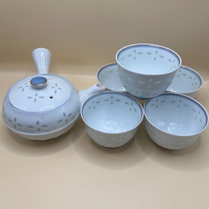 May include: A white ceramic tea set with a teapot and five matching teacups. The teapot has a handle and a lid with a blue accent. The cups and teapot are decorated with a light blue floral pattern and a blue rim.