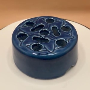 May include: A dark blue ceramic flower frog with a starburst design. The frog has multiple holes for arranging flowers and is sitting on a white surface. The ceramic has a glossy finish.