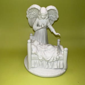 May include: A white ceramic angel figurine with outstretched wings stands over a small bed. The angel is dressed in a long gown and appears to be watching over the person in the bed. The figurine is on a round base.
