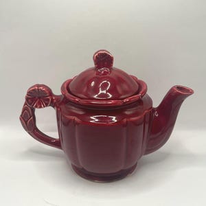 May include: A deep red ceramic teapot with a glossy finish, featuring a scalloped body design, a matching lid with a floral-shaped knob, and a handle adorned with a sculpted flower detail at the top. The spout is curved and tapered.