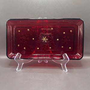 May include: A rectangular ruby red glass tray featuring a decorative pattern of gold-colored snowflakes and small gold dots. The tray has a smooth, glossy finish and is displayed on a clear plastic stand, showcasing its deep red translucent color.