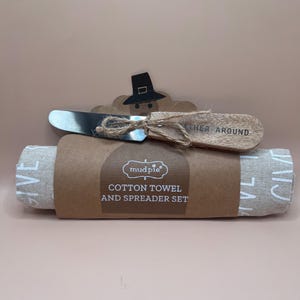 May include: A Mud Pie cotton towel and spreader set featuring a rolled beige towel with the word GIVE printed in white. A metal spreader with a wooden handle is tied to the packaging with twine. The packaging includes a pilgrim hat graphic.