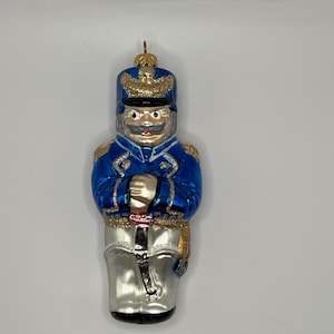May include: A decorative glass ornament shaped like a toy soldier. The soldier wears a blue uniform with gold trim, a matching hat, and silver pants. The ornament has a mustache and is holding a sword. A small metal loop is at the top for hanging.
