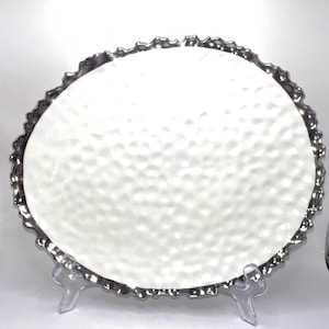 May include: A white, textured, oval-shaped plate with a silver, scalloped edge. The plate is displayed on a clear plastic stand, showcasing its design. The plate's surface has a dimpled texture, adding visual interest to the tableware.