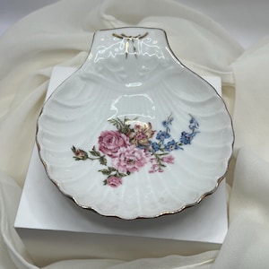 May include: A white porcelain seashell-shaped dish with a gold trim. The dish features a floral design with pink and blue flowers. The dish is likely for decorative purposes.