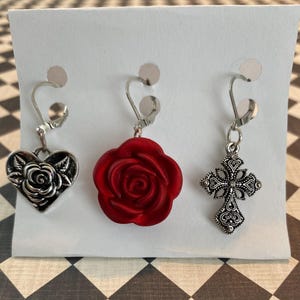 Puede incluir: Tres pendientes de clip en una tarjeta blanca. Los pendientes incluyen un corazón plateado con una rosa, una rosa roja y una cruz plateada con patrones detallados. Estos pendientes están diseñados para orejas no perforadas.