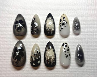 May include: A collection of ten artificial nails with diverse designs. The nails display a palette of black, white, and grey. Some nails feature floral patterns, while others have speckled or textured designs. The nails are oval-shaped.