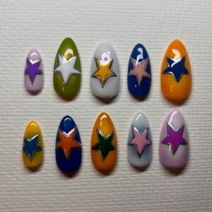 May include: A set of ten artificial nails in various colors, including pink, green, white, blue, and orange. Each nail features a star design with a silver outline. The nails are oval-shaped and arranged in a neat row.