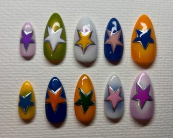 Puede incluir: Un conjunto de diez uñas artificiales en varios colores, incluyendo rosa, verde, blanco, azul y naranja. Cada uña presenta un diseño de estrella con contorno plateado. Las uñas son ovaladas y están dispuestas en fila.