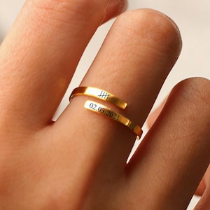Può includere: Un anello avvolgente color oro con incisioni e la data 02.03.2021. L'anello è su un dito e il design include una serie di linee verticali e la data.