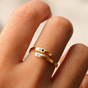 Puede incluir: Un anillo envolvente de color dorado con texto grabado y un símbolo de corazón. El anillo está en un dedo, y el texto incluye iniciales y una fecha. El diseño del anillo es una banda continua que se envuelve alrededor del dedo.
