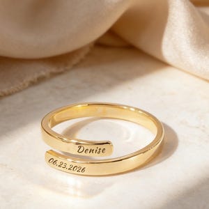 May include: A gold-coloured, adjustable ring engraved with the name "Denise" and the date "06.23.2026". The ring features a spiral design and is shown on a light surface, with a soft, neutral-toned fabric in the background.