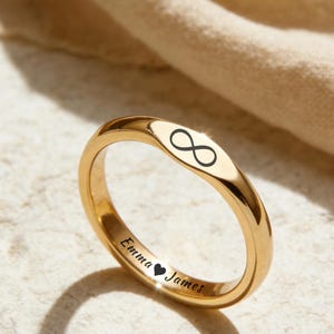 May include: A gold ring featuring an infinity symbol and the engraved text "Emma ♥ James." The ring is displayed on a neutral surface. The ring's design is simple and elegant, with a polished finish.