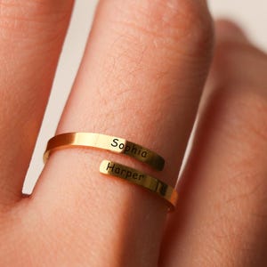 Puede incluir: Un anillo envolvente de color dorado con los nombres "Sophia" y "Harper" grabados. El anillo se muestra en un dedo, con los nombres en la parte delantera.