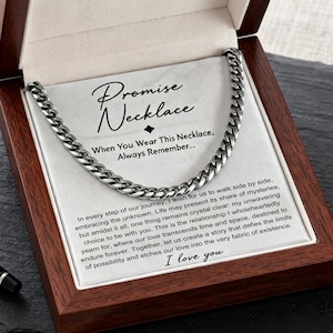 May include: A silver-toned curb chain necklace displayed inside an open wooden gift box. The box contains a white card with the text Promise Necklace and a printed message. Two black rings and a black pen are positioned outside the box.