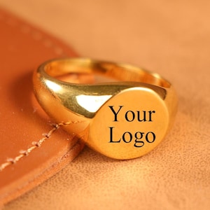 以下が含まれることがあります： 丸みを帯びた顔の金色のシグネットリング。リングの表面には「Your Logo」の文字がプリントされています。リングは茶色の革の表面に置かれています。