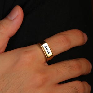 May include: A gold-colored ring with the name "Adam" engraved on the front. The ring is rectangular and worn on a finger. The background is a dark, solid color.